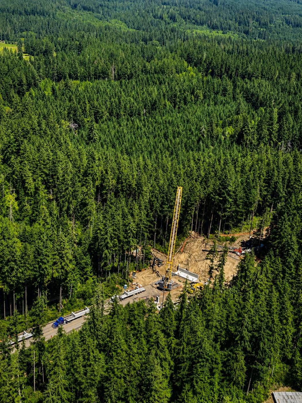 Oblique aerial photo taken from Harbor View Air's Cessna 172 N3843R of a construction site with a tall yellow crane in a clearing surrounded by dense evergreen forest, beside a road lined with trucks