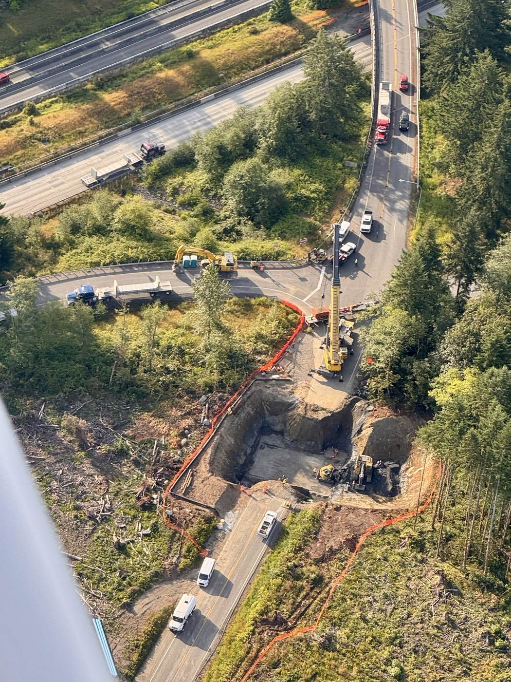 Oblique aerial photo taken from Harbor View Air's Cessna 172 N3843R, wing strut visible at lower left, of a large excavation pit with a crane and orange safety fencing next to a highway interchange