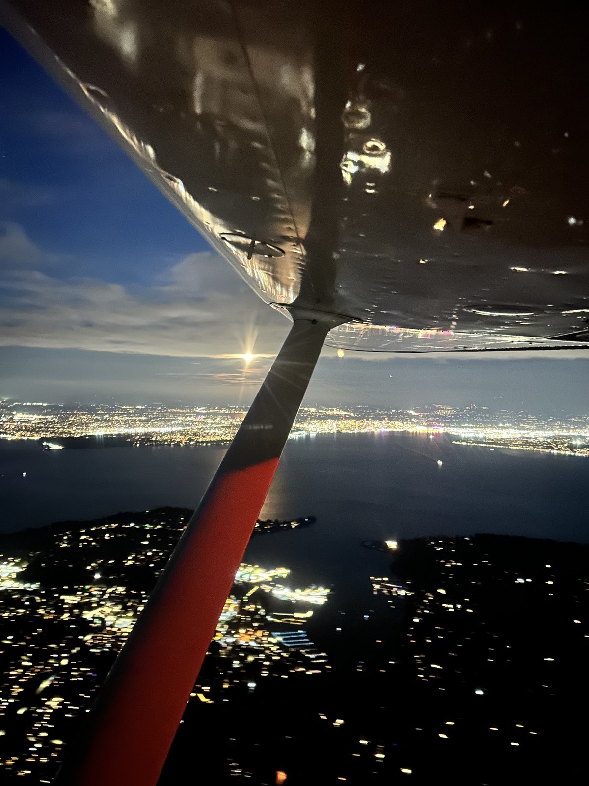 Evening view from the Cessna 172 N3843R in flight, with the red-striped wing strut in the foreground and city lights along the water below a bright moon