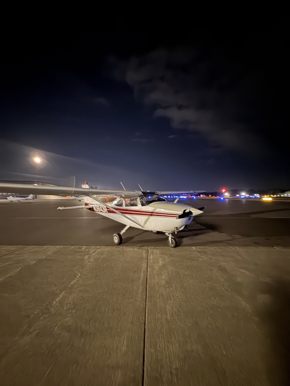 White Cessna 172 N3843R with a maroon stripe parked on a lit airport ramp at night, with the moon low in the sky