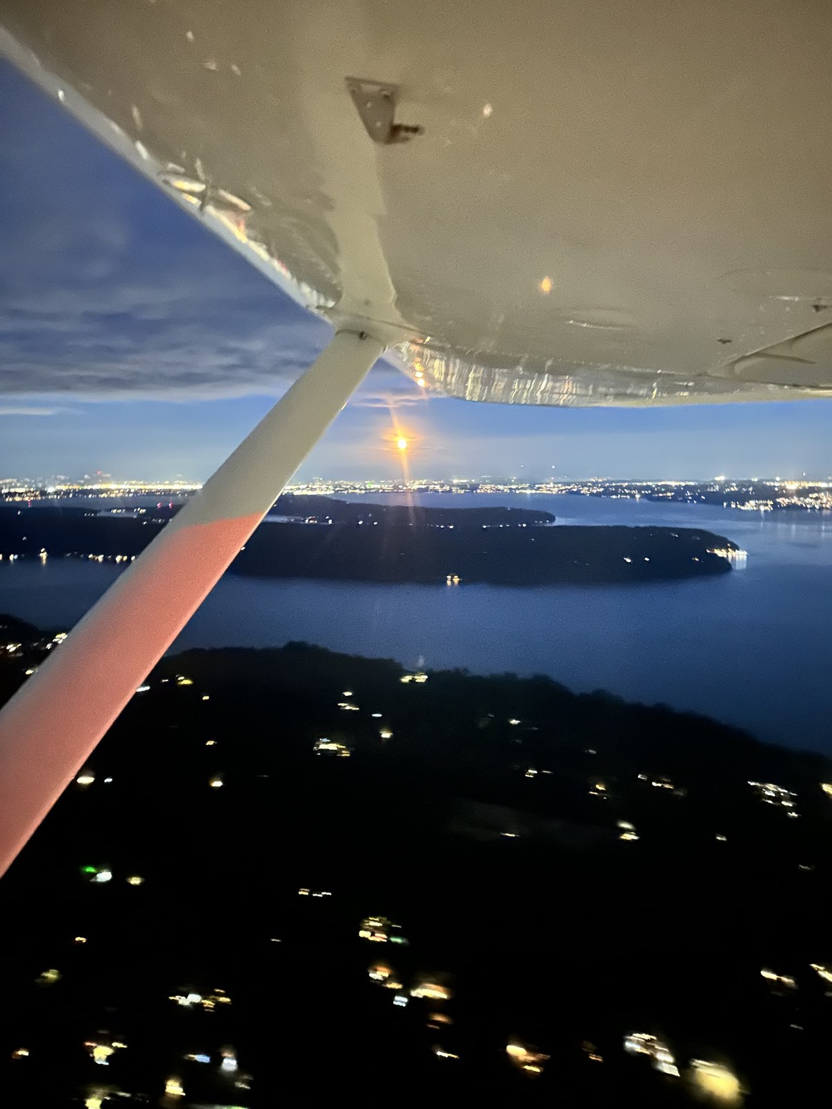 View from inside the Cessna 172 N3843R in flight at dusk, looking under the white wing and its strut toward water, a forested shoreline, and distant lights